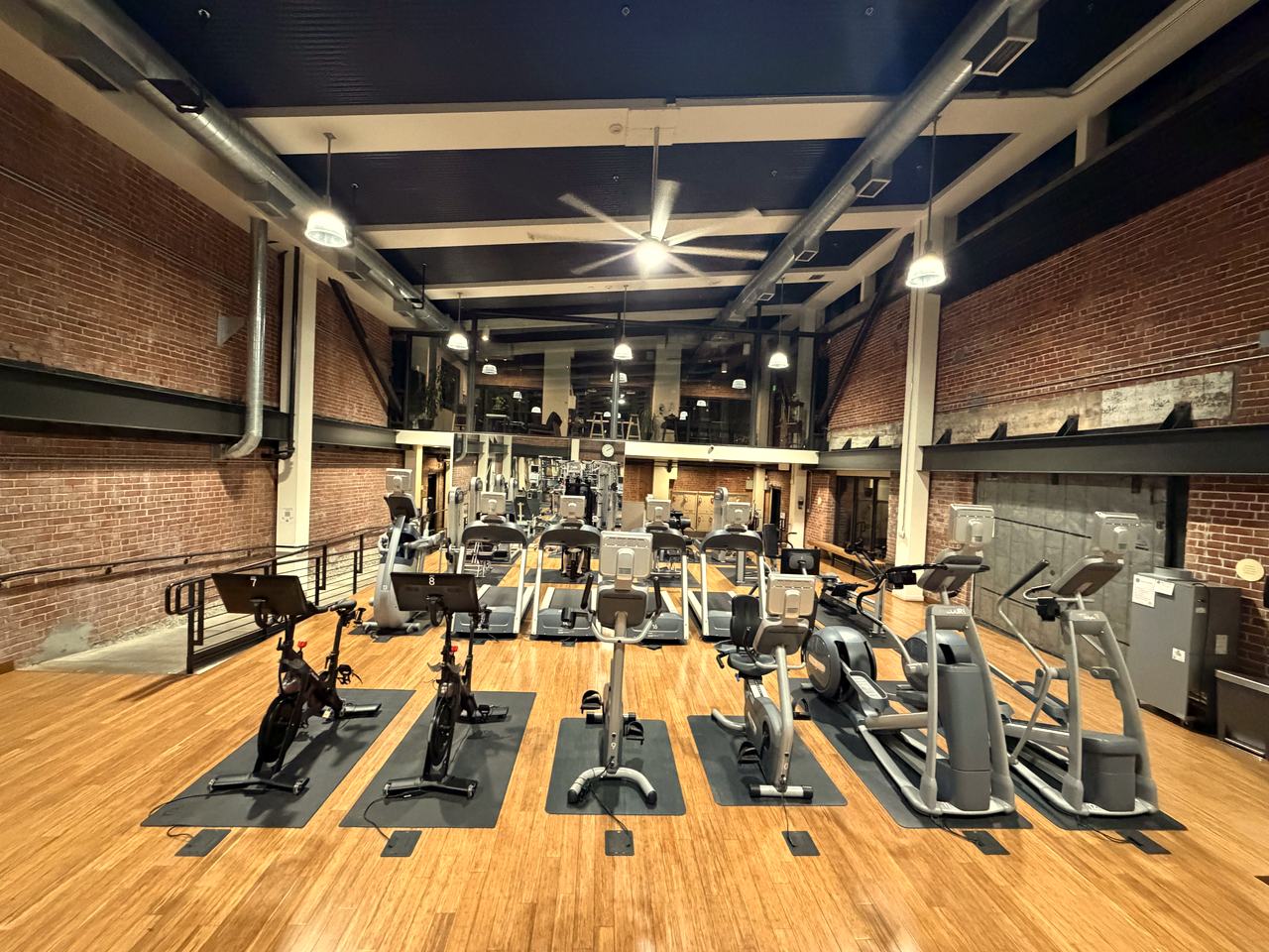 Inside the Boiler Room fitness center — cardio equipment on bamboo floors beneath the original brick walls and steel beams, with the Mezzanine Lounge glass above